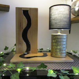 Création artisanale composée d'une planche en bois lasuré intégrant une rivière sculptée en résine noire brillante, accompagnée d'une lampe cylindrique dorée avec abat-jour noir.