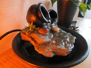 Fontaine d'intérieur artisanale noire nommée Le Murmure des Jarres, composée de deux petites jarres sur une pierre avec un écoulement d'eau lumineux.